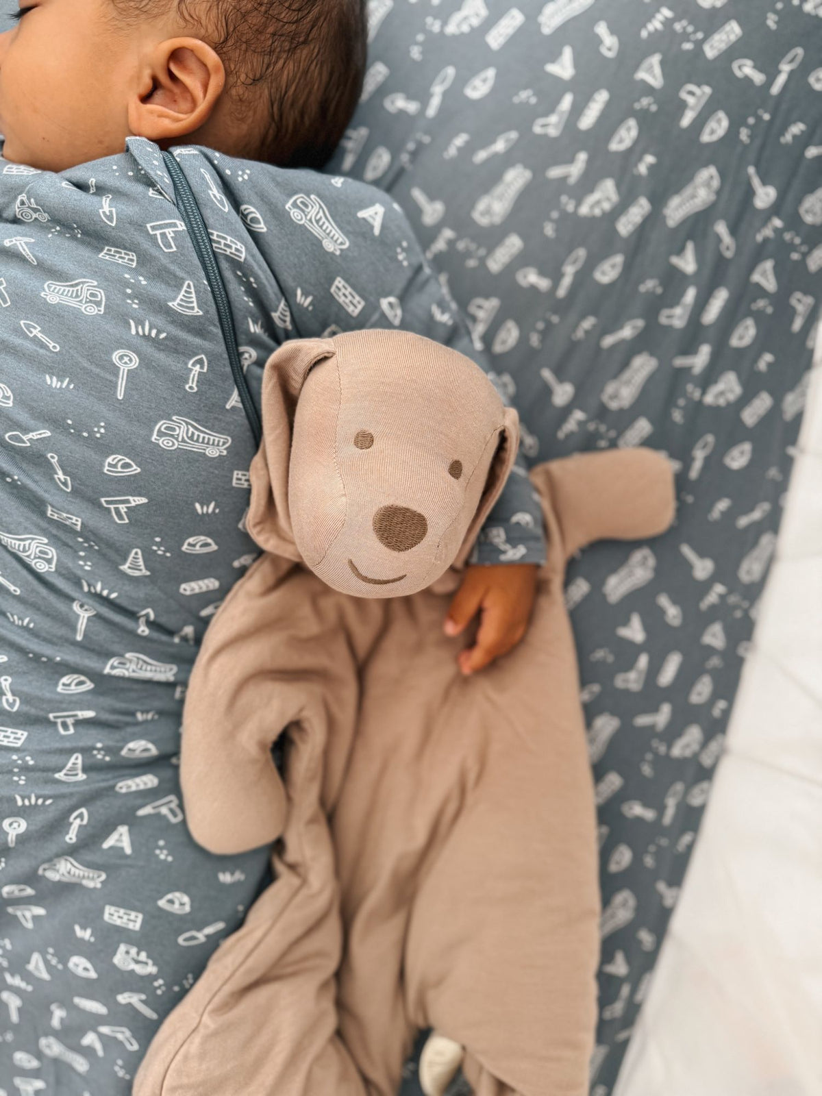 Taupe dog plush lovey with smiling face held against a blue patterned onesie.