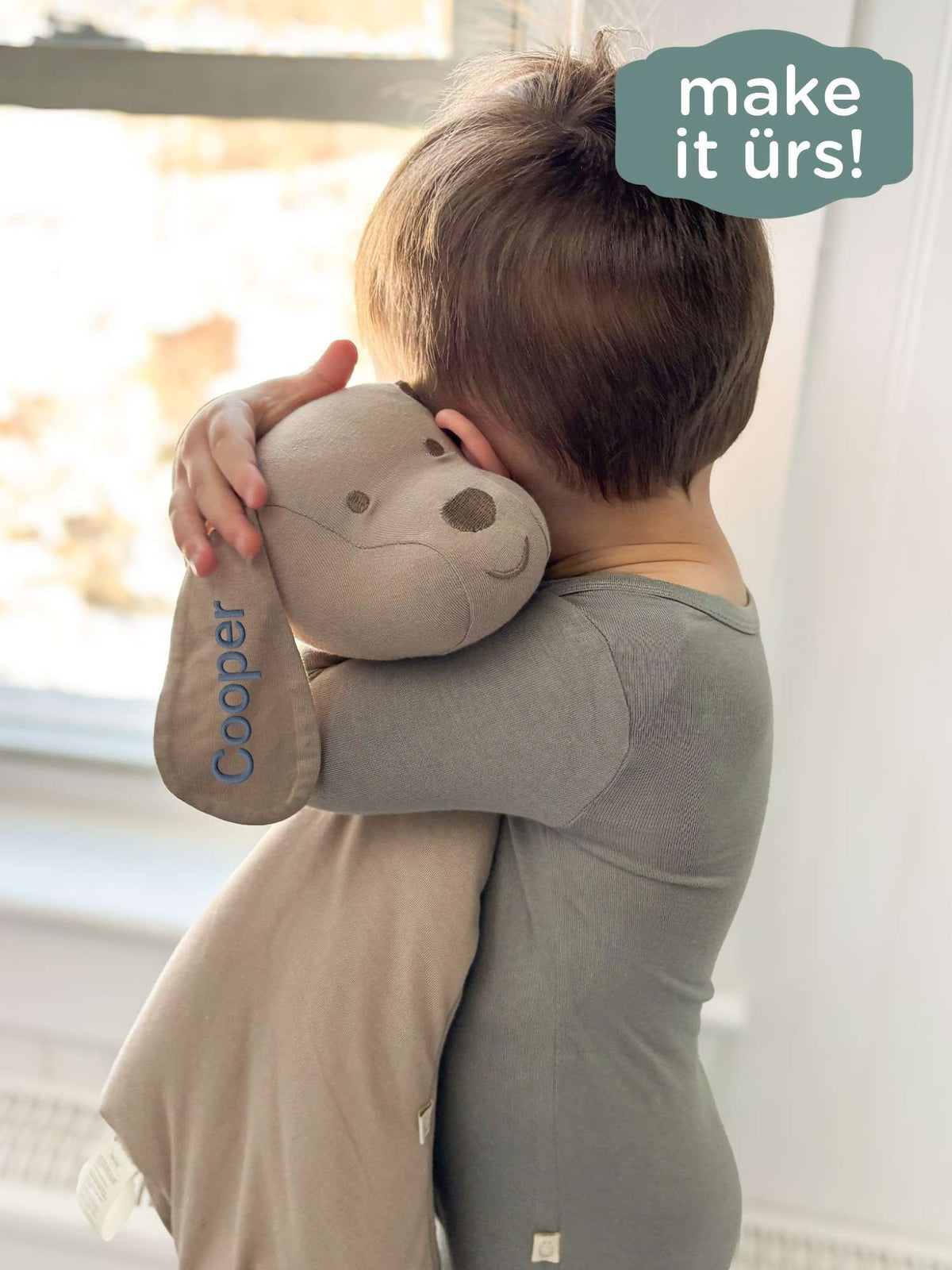 Taupe puppy plush lovey hugged close by a baby near a window.