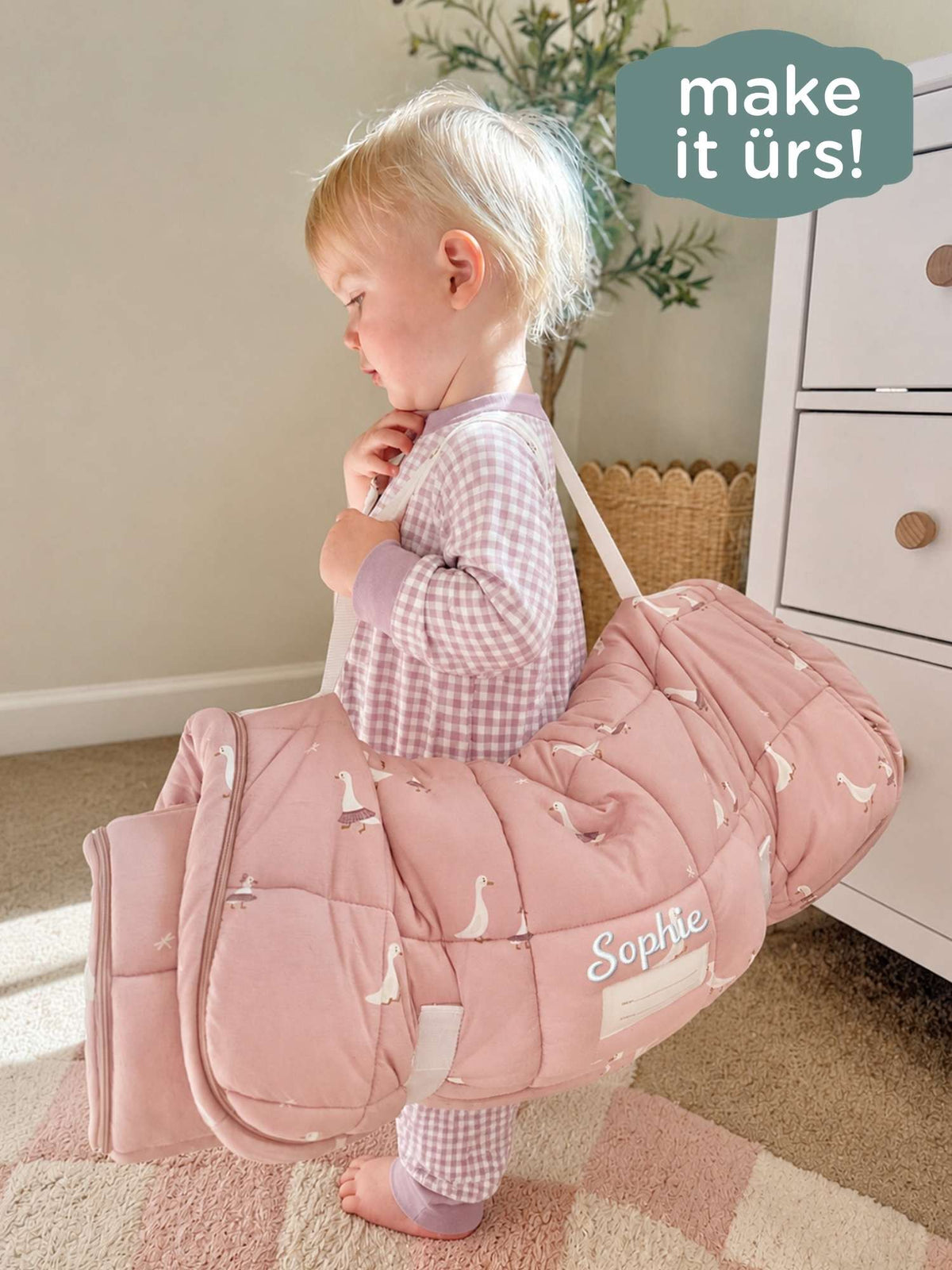 Light pink nap mat with white goose pattern, baby standing beside the folded mat