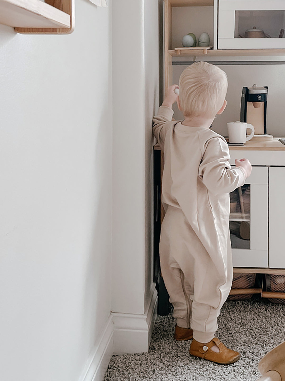 Blush fleece baby jumpsuit with long sleeves and cuffs, standing and looking toward kitchen