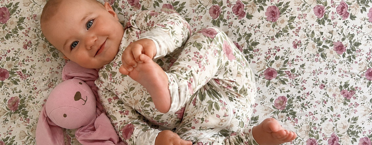 A smiling baby lying on a floral-patterned blanket, wearing a matching long-sleeve footless onesie and holding a pink plush toy.