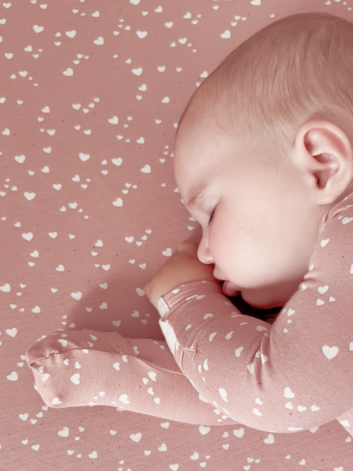 Dusty rose crib sheet with small heart pattern, tucked under baby in side view