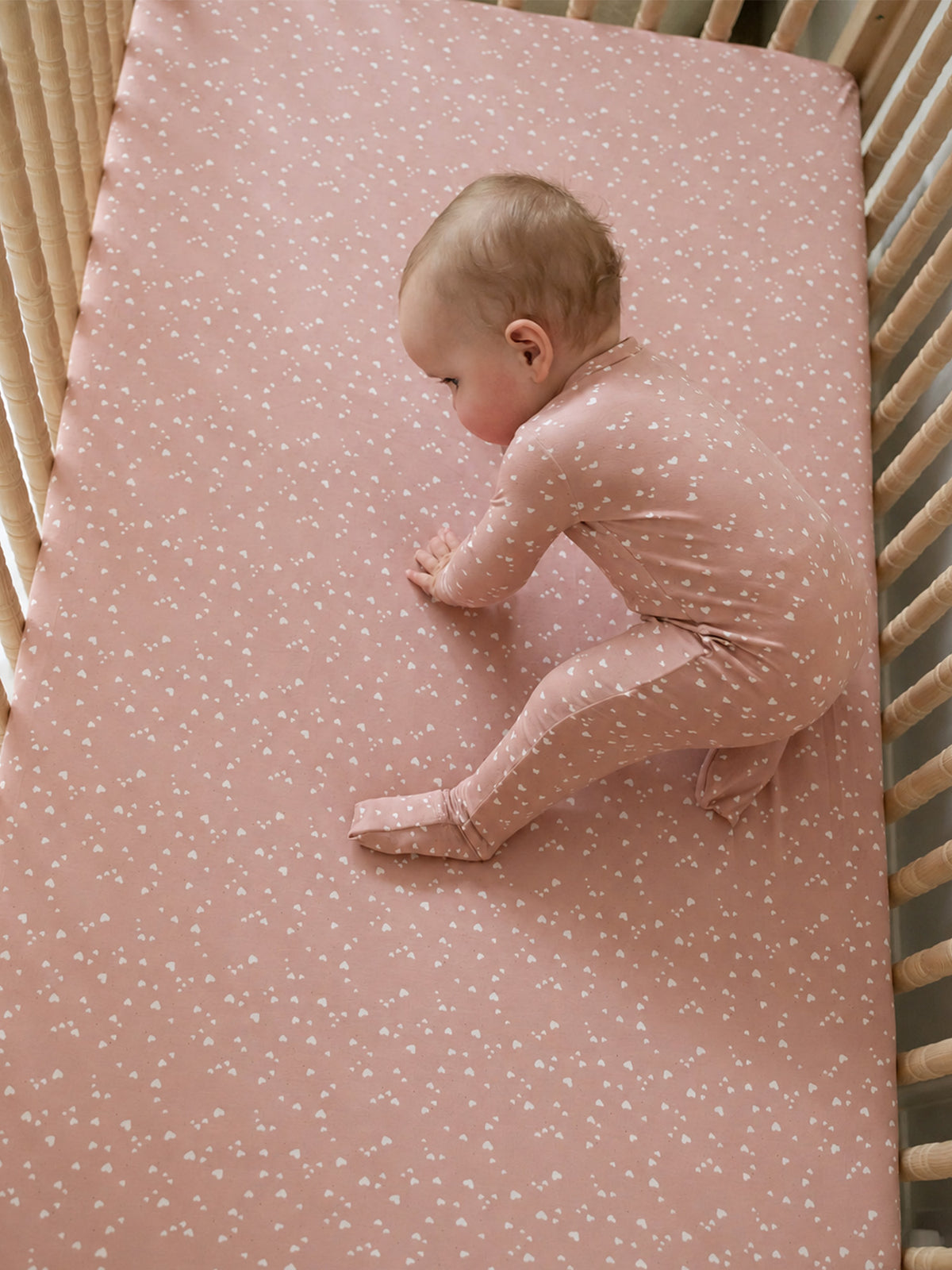 Dusty rose crib sheet with tiny heart print, baby lying and stretching on sheet