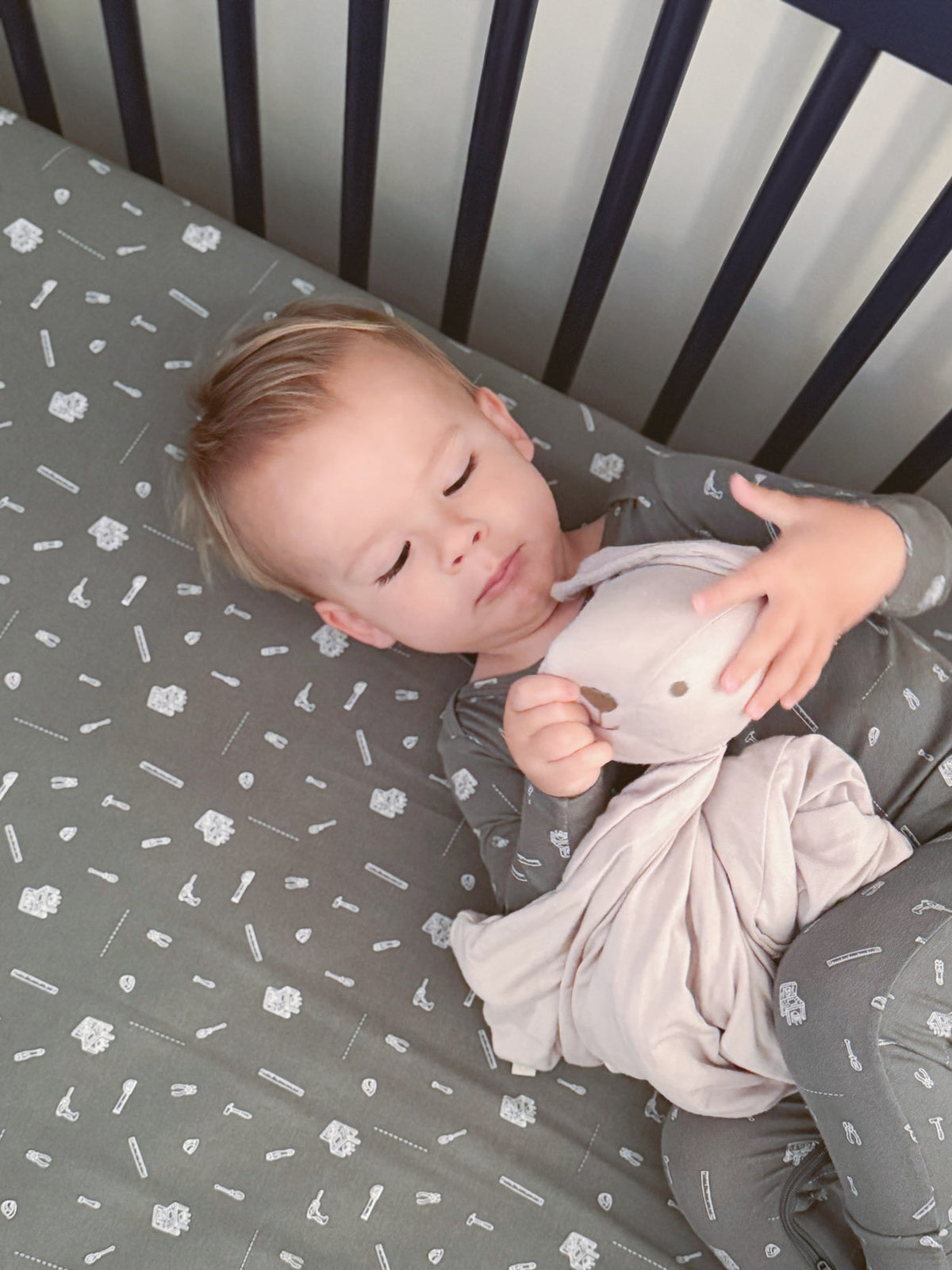 Grey crib sheet with white tool icons, baby lying and holding a plush