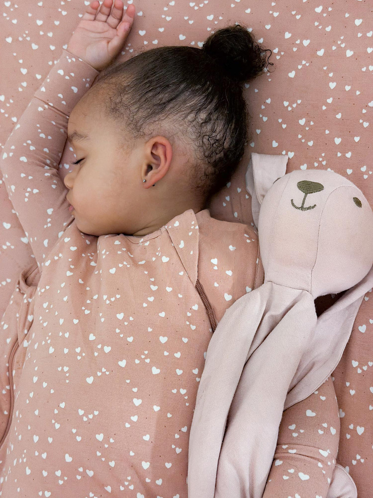 Pink baby sleep sack with heart print, long sleeves and zipper front beside plush toy