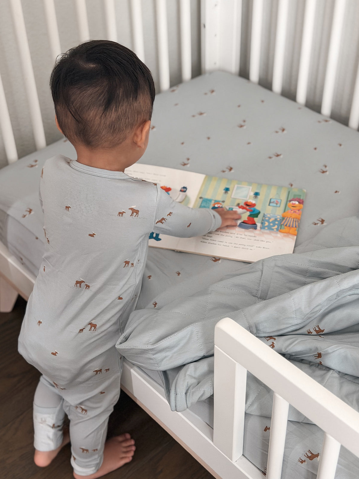 Light gray baby blanket with moose print on a cot, toddler reading beside it.
