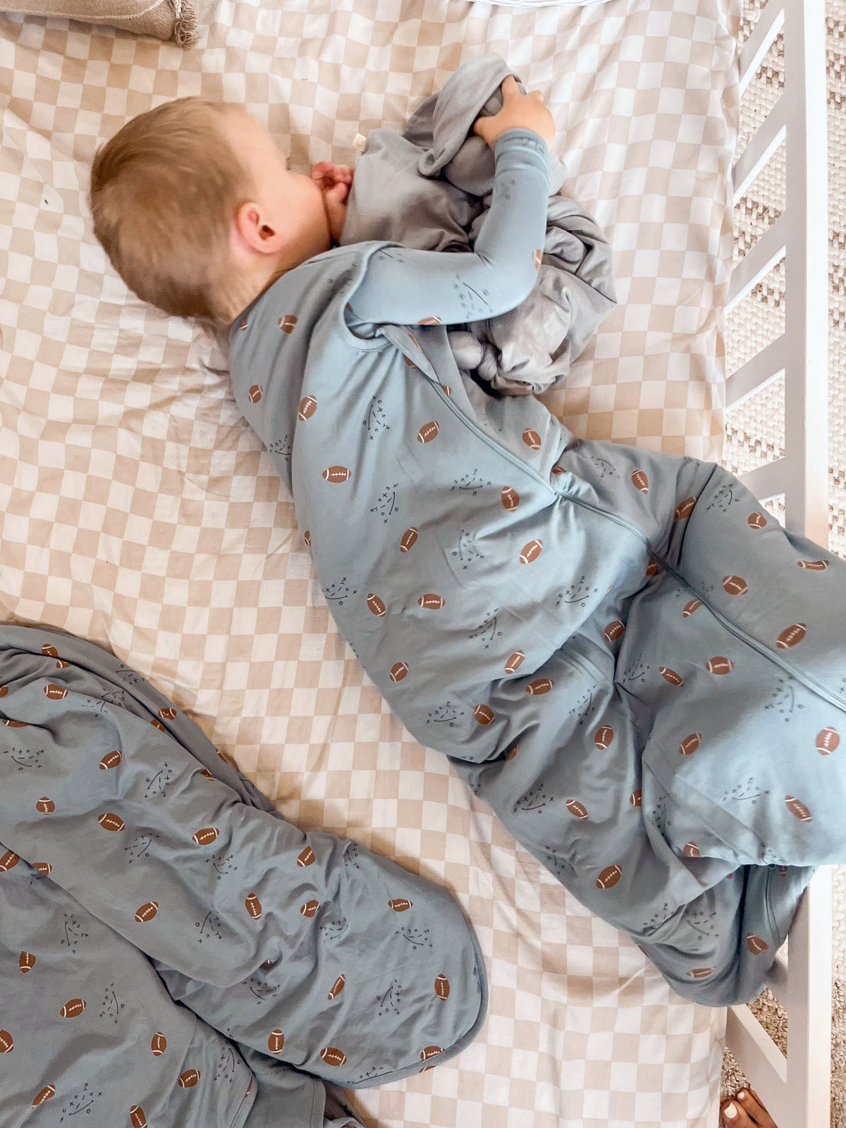 Light blue football-print sleep sack with long sleeves and zipper, toddler lying on bed