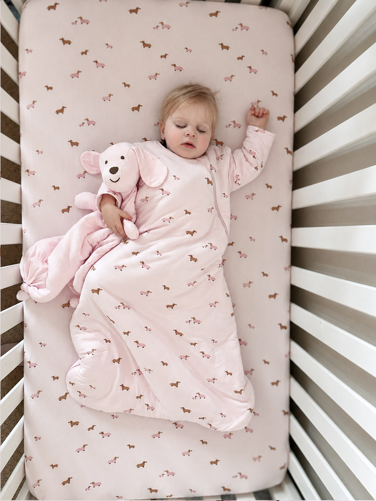 Crib sheet with allover dog print in pink and tan, with baby lying on it