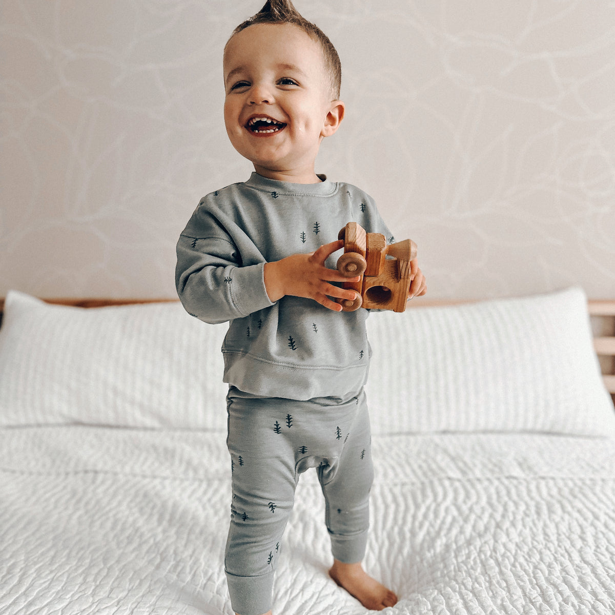 toddler boy standing on white bedsheets, holding a wooden toy train in a fleece jogsuit with diaper zipper
