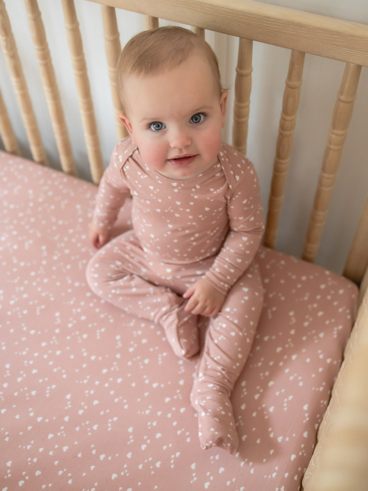 Dusty rose crib sheet with tiny heart print, baby lying on it in crib