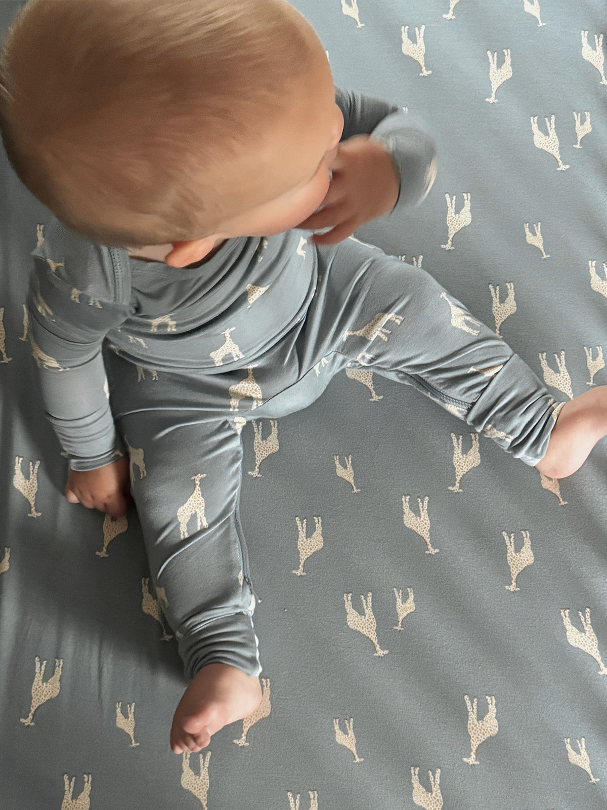 Blue-gray footed pajamas with beige giraffe print, baby sitting and leaning forward