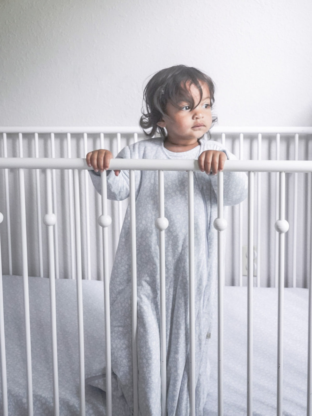 Light gray patterned long-sleeve sleep sack with zipper closure, worn and standing in crib.